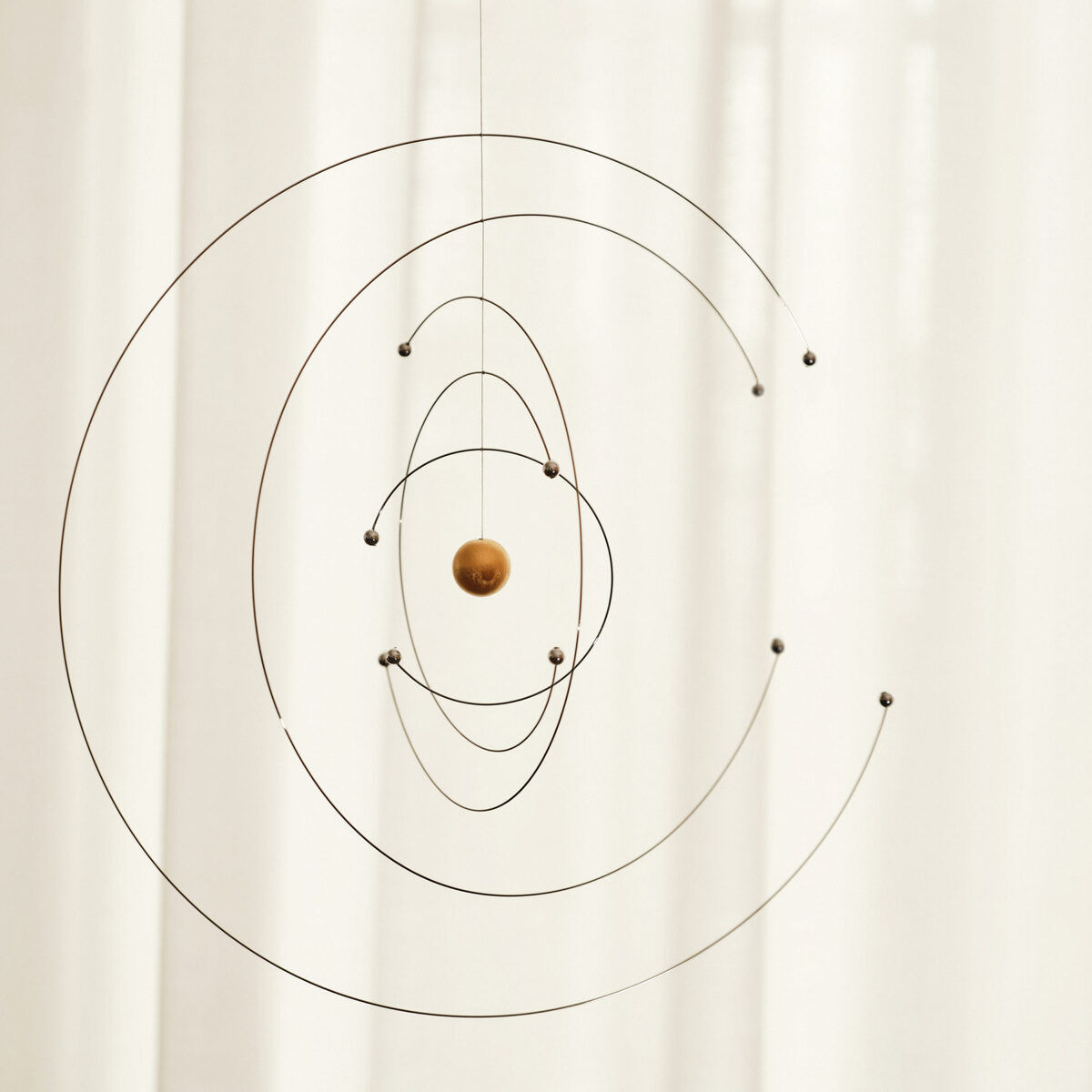 Ein wissenschaftliches Mobile namens »Niels Bohr« bestehend aus konzentrischen Metallringen mit kleinen Holzkugeln, die Planeten darstellen. Es hängt vor einem hellen, weißen Hintergrund.