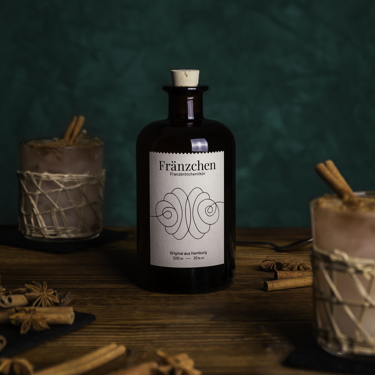 Eine dunkelbraune Flasche des Likörs »Fränzchen« steht auf einem Tisch, umgeben von zwei Cocktailgläsern, die mit Zimtstangen dekoriert sind. Der Hintergrund ist dunkelgrün und die Tischplatte hat eine rustikale Holzoptik.
