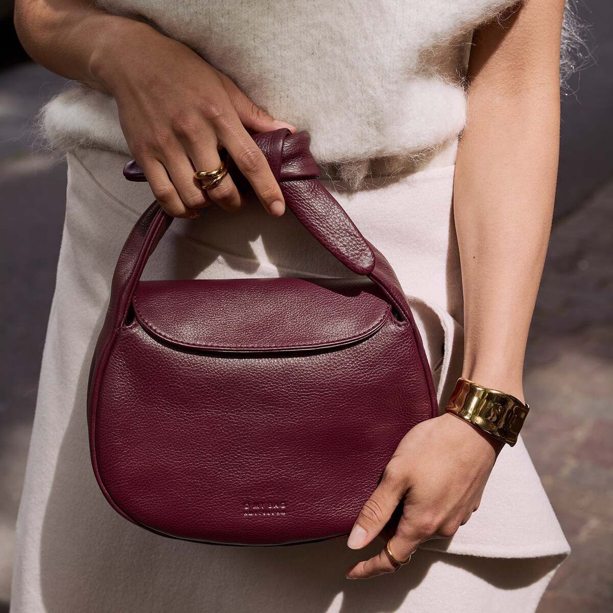 Eine Frau hält eine bordeauxrote Lederhandtasche mit elegantem Henkel. Sie trägt ein cremefarbenes, ärmelloses Oberteil und einen goldenen Armreif. Im Hintergrund ist ein unscharfer Außenbereich zu sehen.