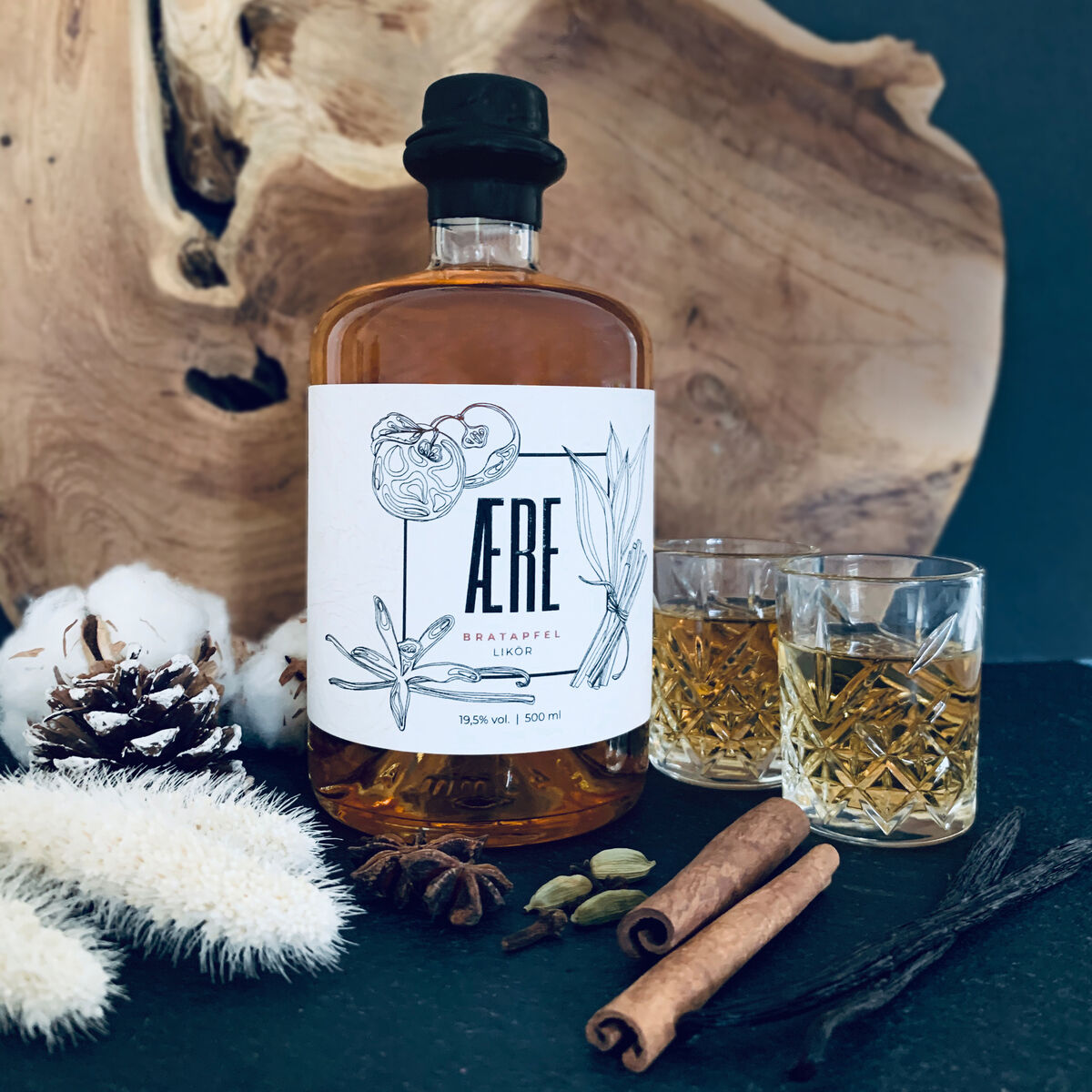 Eine Flasche Aere Bratapfellikör steht auf einem Tisch vor einem Holzbrett. Um die Flasche herum liegen Zimtstangen, Vanilleschoten, Anis, Kardamom und weiße Dekorationselemente. Daneben stehen zwei gefüllte Gläser.