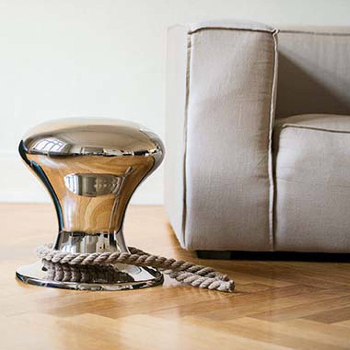 Ein moderner Hocker namens 'Hamburger Hocker mit Segeltau' steht neben einem beige-farbenen Sofa auf einem Holzboden. Der Hocker besteht aus glänzendem Metall und ist mit einem dicken Segeltau verziert, das um den Sockel gewickelt ist. Der Raum wirkt hell und modern.