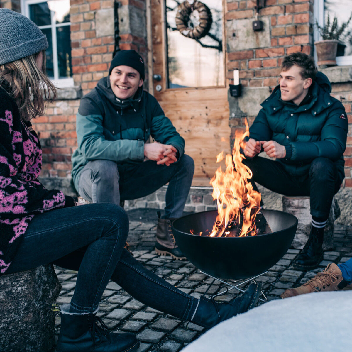 Drei Personen in Winterkleidung sitzen um eine Feuerschale und wärmen sich an einem Lagerfeuer, vor einem Backsteinhaus im Freien.