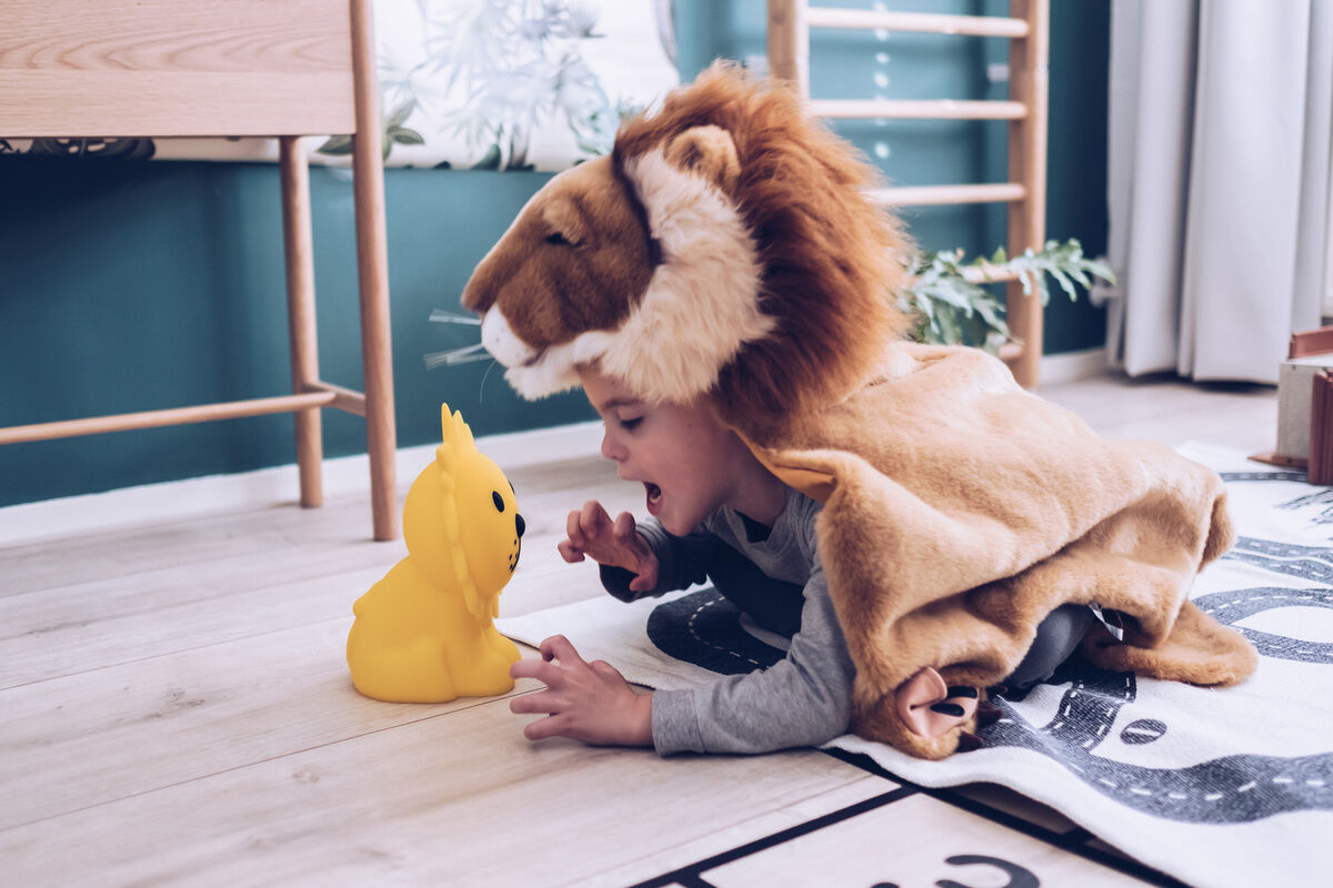 Ein Kind, verkleidet in einem Löwenkostüm, spielt mit einem gelben Nachtlicht in Form eines Löwen auf dem Boden eines Kinderzimmers.