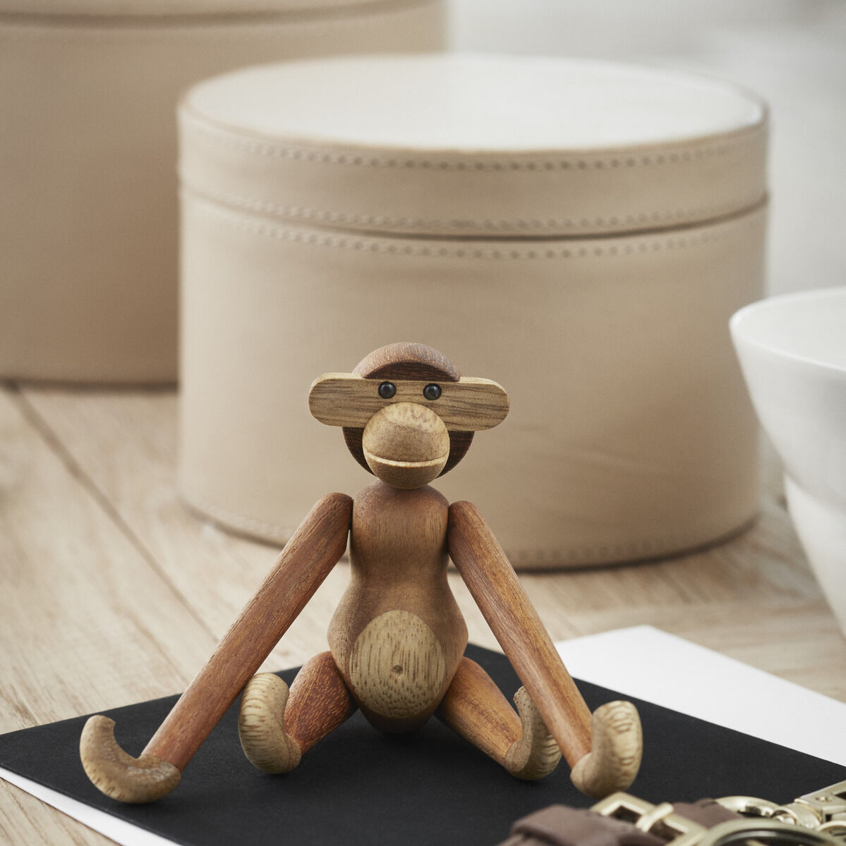 Holzfigur eines Affen von Kay Bojesen, platziert auf einem dunklen Untergrund und umgeben von verschiedenen Alltagsgegenständen wie Schlüssel und Dekorationsobjekten.
