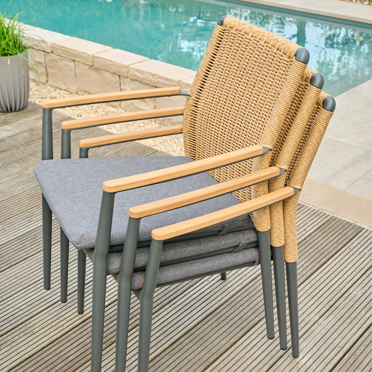 Ein Stapelsessel »Roma« aus einem robusten, wetterfesten Materialmix, bestehend aus Rattan und Holzoptik. Der Stuhl ist bequem und modern, in beige und grau Tönen gehalten, und steht auf einem Holzdeck neben einem Pool.