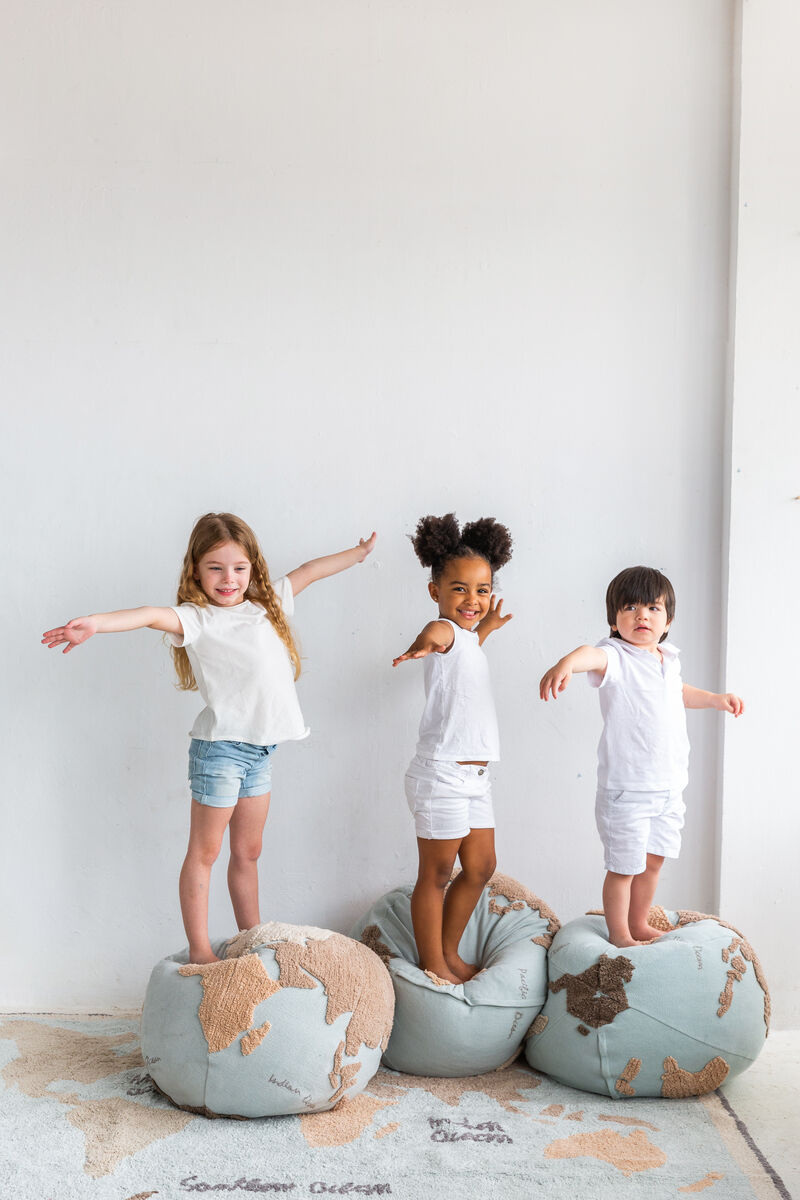 Drei Kinder stehen auf Sitzkissen mit Weltkartendesign, lächelnd und die Arme ausbreitend vor einer weißen Wand.
