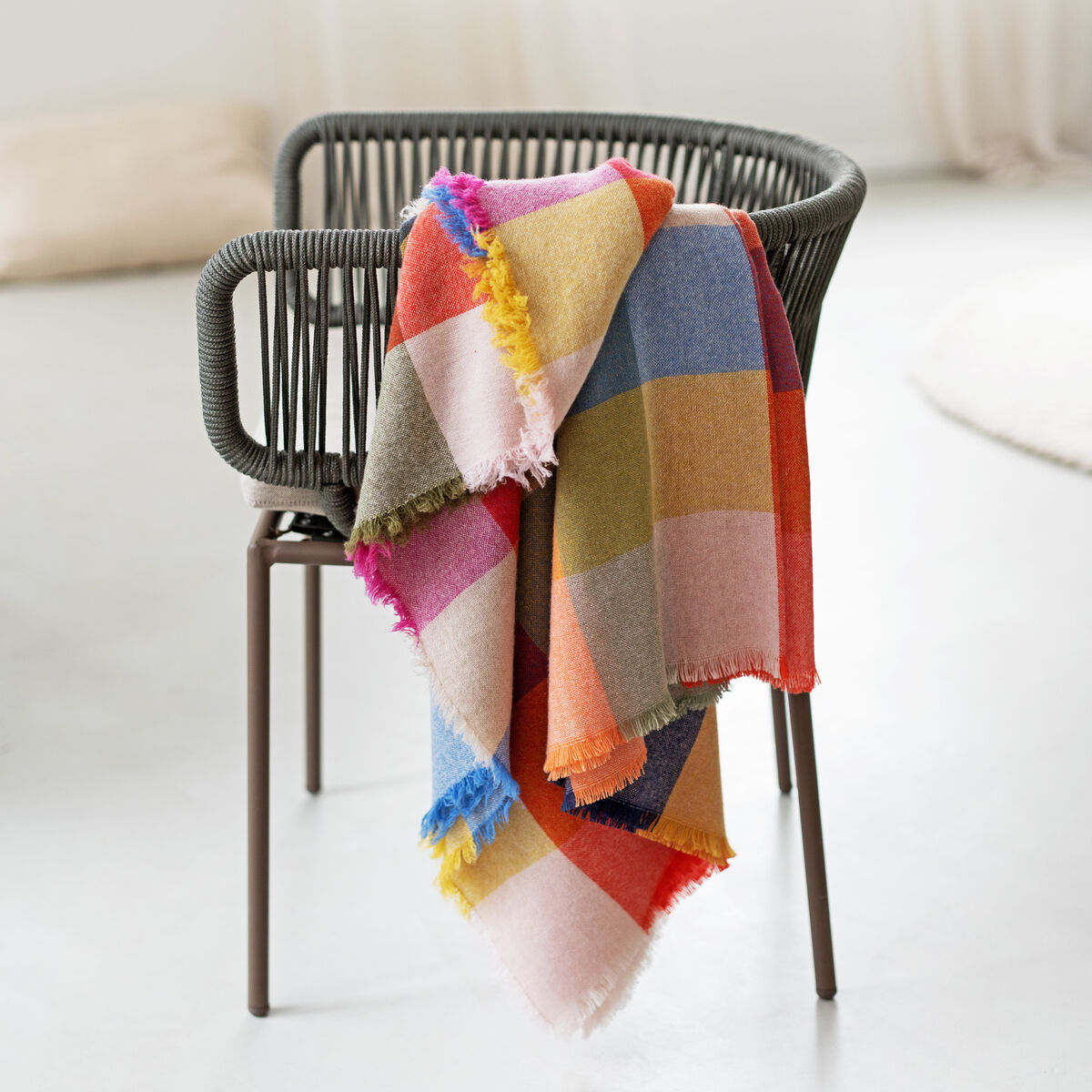 Eine luxuriöse Merino-Kaschmir-Decke »St. Tropez« mit großem, farbenfrohem Karomuster liegt locker über einen modernen Stuhl mit dunklem Metallgestell und geflochtener Rückenlehne in einem hellen, minimalistischen Raum mit weißen Wänden und sanften Lichtverhältnissen.