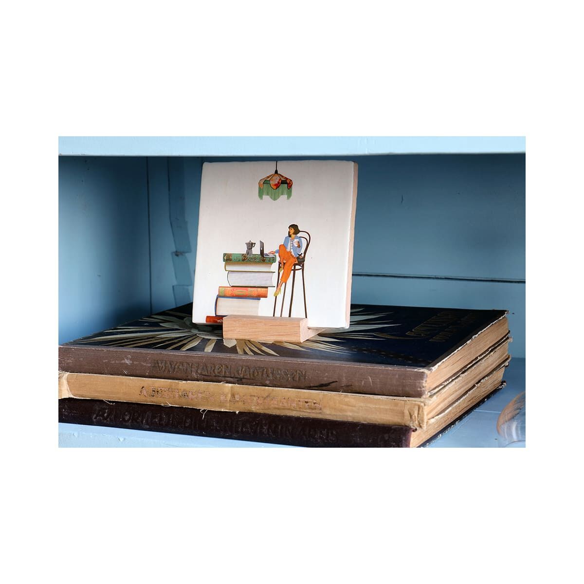 Keramikfliese mit illustrativem Aufdruck, die eine Frau an einem Küchentresen unter einer Lampe zeigt. Die Fliese steht auf einem Holzständer und ist auf einem Stapel alter Bücher in einem hellblauen Regal arrangiert. Insgesamt entsteht ein wohnliches, kreatives und humorvolles Ambiente.