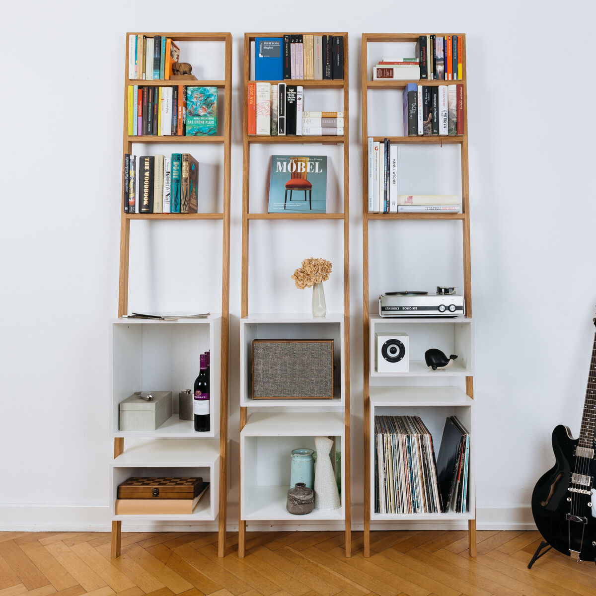 Ein modernes Leiterregal aus Holz und weißen Elementen, gefüllt mit Büchern, Dekorobjekten und elektronischen Geräten, steht an einer weißen Wand. Ein schwarzes Musikinstrument lehnt an der rechten Seite.