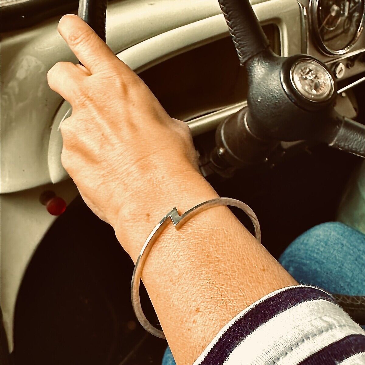 Hand am Lenkrad eines Oldtimers, am Handgelenk ein runder silberner Armreif, Sonderedition »ZEIT« von HML Berlin. Das Z-Design ist klar erkennbar. Person trägt gestreiftes Oberteil. Lenkrad und Armaturenbrett im Hintergrund, Retro-Stimmung.
