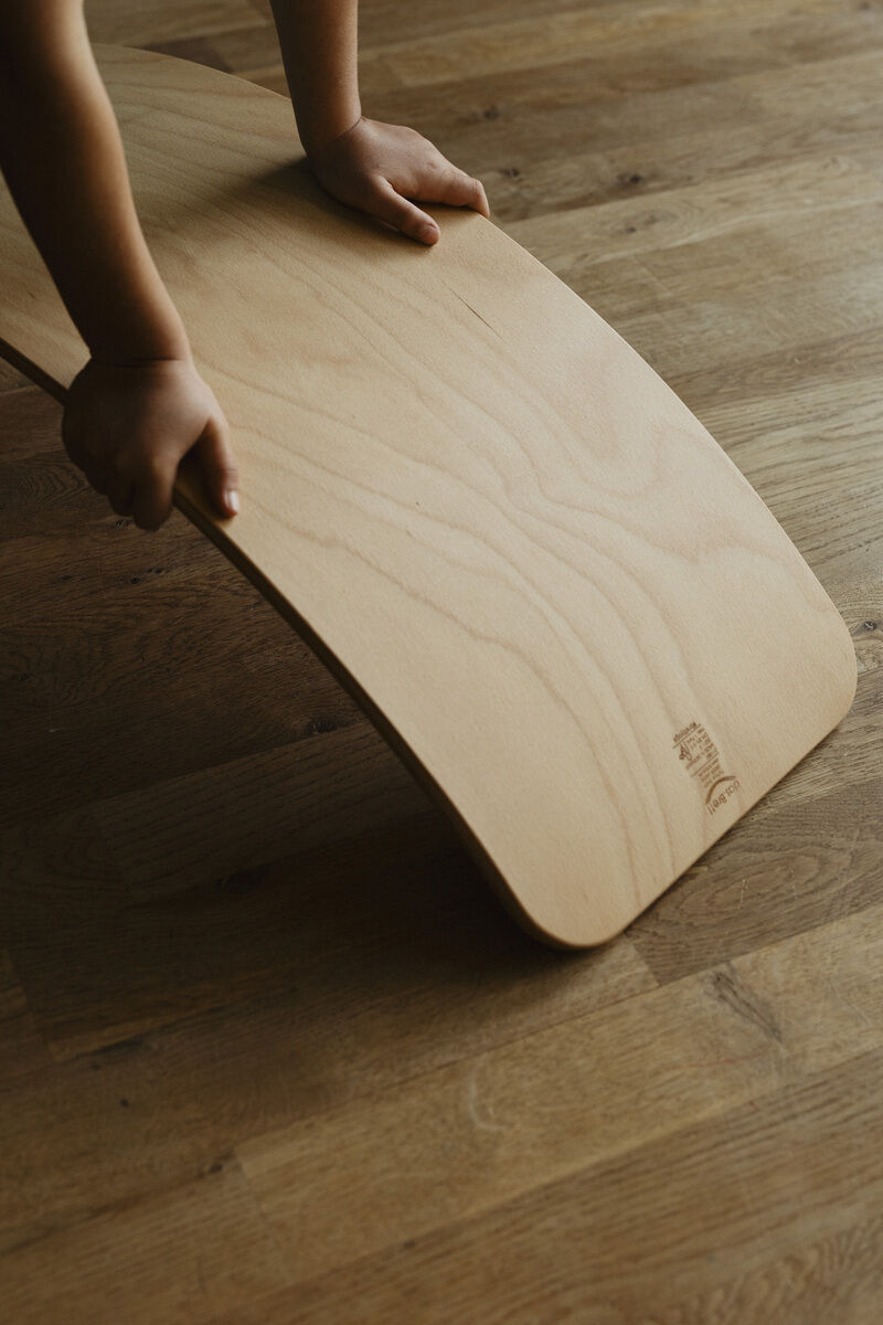 Holz-Balance-Board mit geölter Oberfläche, auf einem Holzboden stehend, mit Kinderhänden, die das Brett halten.