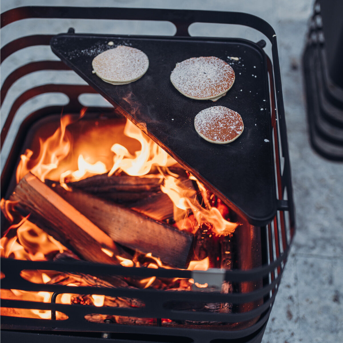 Eine moderne Feuerstelle mit einer integrierten Grillplatte (Plancha). Auf der Platte brutzeln drei Pfannkuchen, während unter dem Grillholz lodernde Flammen sichtbar sind. Outdoor-Kochsituation bei kaltem Wetter.