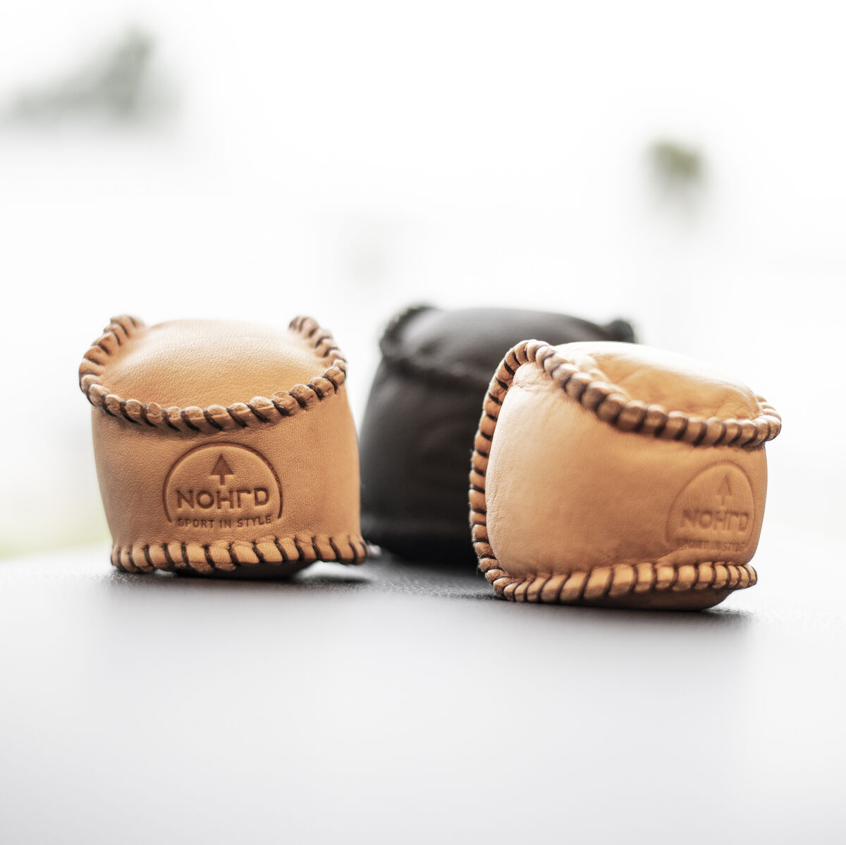 Drei HaptikBälle aus Leder in Naturfarben auf einer glatten Oberfläche. Zwei Bälle sind hellbraun, einer ist dunkelbraun. Die Bälle haben handgenähte Nähte und das Logo von NOHrD.