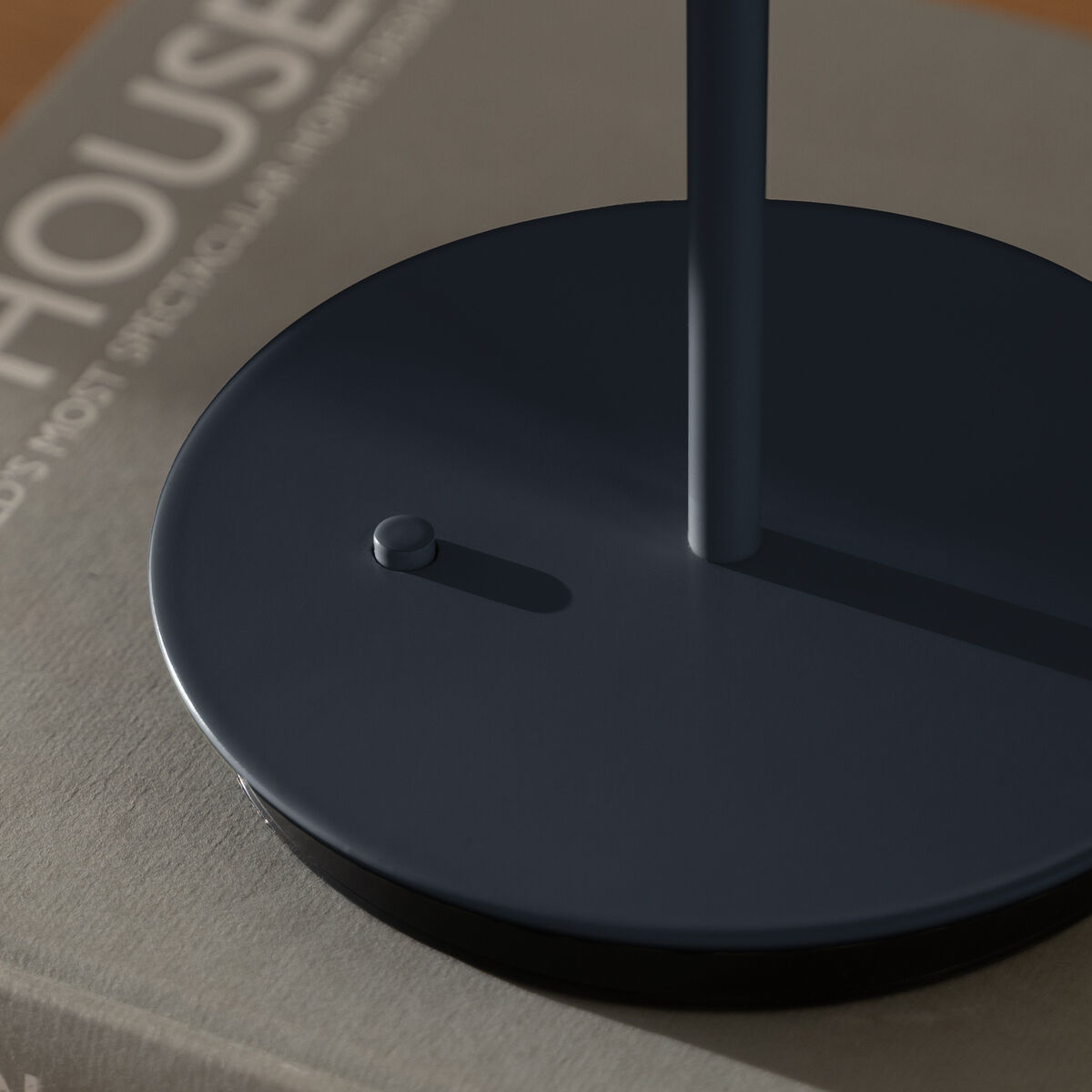 Die Abbildung zeigt den Fuß einer eleganten, kabellosen Tischlampe der ZEIT Sonderedition Asteria Move in Midnight Blue auf einem neutralen Buch mit hellem Einband auf einer Holzoberfläche. Das minimalistische und moderne Design hebt sich durch die matte dunkelblaue Farbe und die schlichte Form hervor. Die Szene vermittelt einen stilvollen und ruhigen Einrichtungsstil.