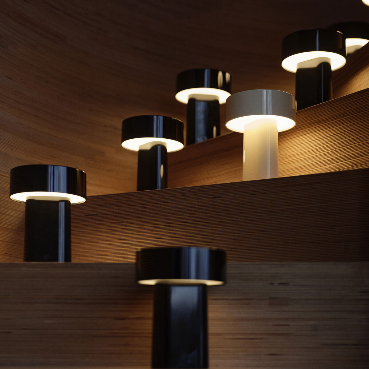 Mehrere tragbare Lampen der Serie Oblong stehen erleuchtet auf hölzernen Treppenstufen in einem modernen Innenraum. Die Lampen haben ein minimalistisches, zylindrisches Design in Schwarz und Weiß und erzeugen ein sanftes, warmweißes Licht, das stimmungsvolle Akzente im Raum setzt.