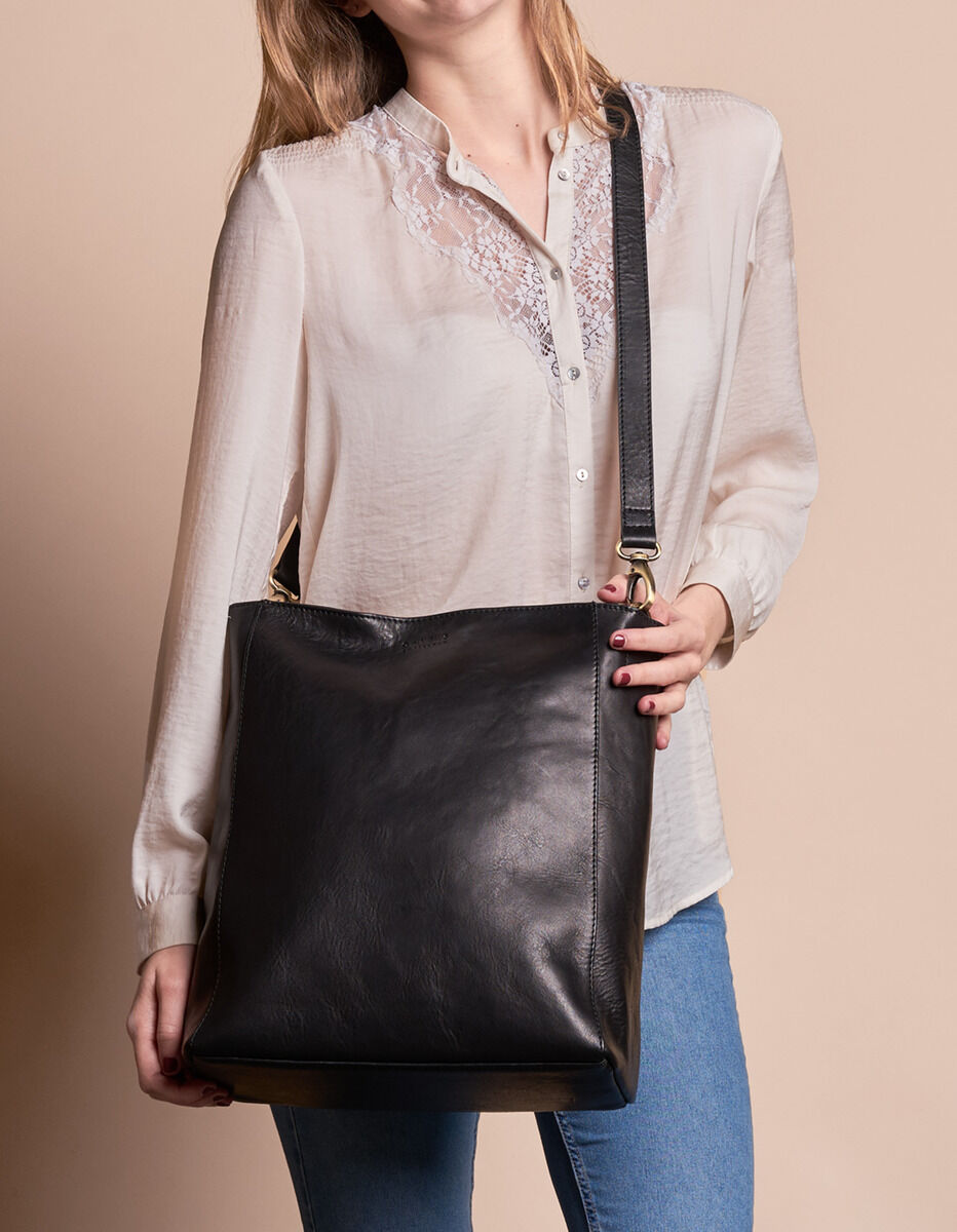 Eine Frau trägt eine schwarze Ledertasche über der Schulter. Sie trägt eine beige Bluse und blaue Jeans.