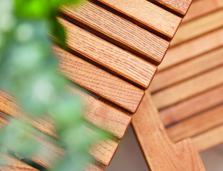 Nahaufnahme eines Holztisches mit klaren und parallelen Holzlatten. Auf der linken Seite ist eine unscharfe grüne Pflanze sichtbar.