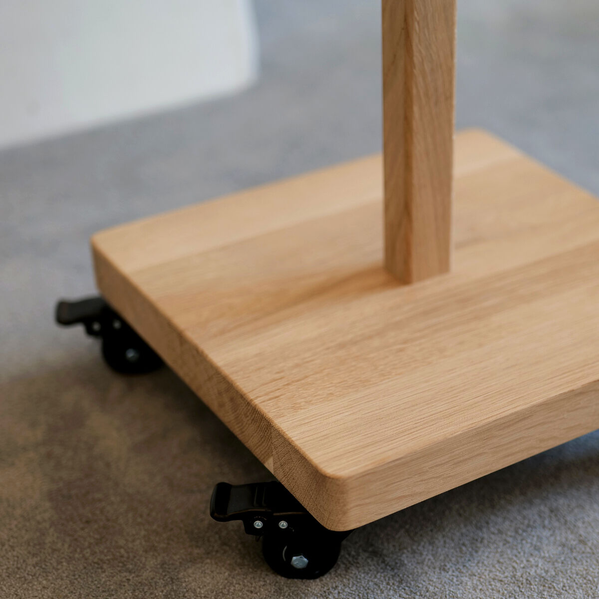 Mobiles Device Stativ "ERA" von Frau Caze aus naturfarbener Eiche und Birkensperrholz mit rechteckigem, massivem Standfuß und schwarzen Lenkrollen auf grauem Teppichboden. Das Design ist schlicht, modern und hochwertig verarbeitet.