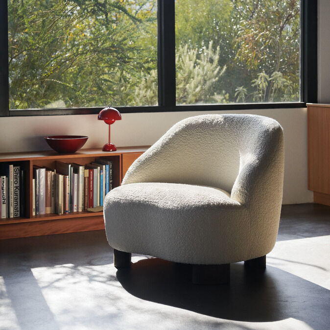 In einem modern eingerichteten Raum steht ein cremefarbener, gepolsterter Designer-Sessel auf grauem Boden vor einem großen Fenster mit Blick ins Grüne. Links von ihm steht eine rote Flowerpot VP9 Akkutischleuchte von &Tradition auf einem niedrigen Regal mit Büchern und einer roten Schale.