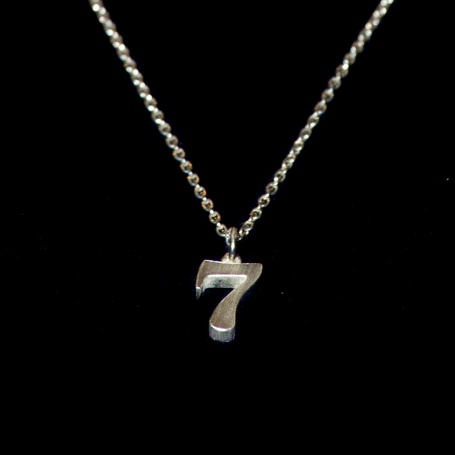 Detailreiche Aufnahme eines silbernen Anhängers in Form der Zahl 7 an einer feinen Kette vor schwarzem Hintergrund. Der Anhänger wirkt massiv und hochwertig verarbeitet, das matte 925er Silber reflektiert dezent das Licht.