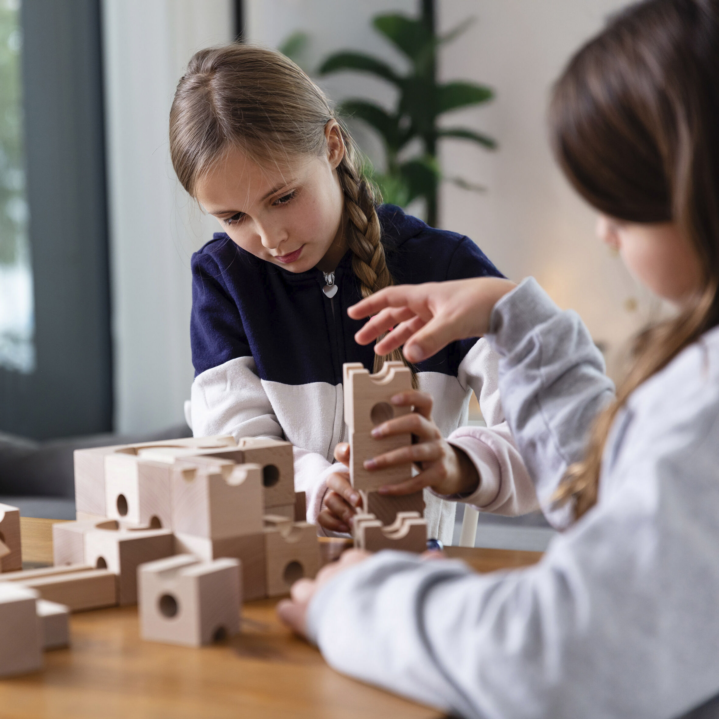 Zwei Kinder spielen mit Holzbausteinen auf einem Tisch in einem hellen Raum. Ein Mädchen stapelt konzentriert die Blöcke, während ein weiteres Kind die Szene beobachtet. Im Hintergrund ist eine grüne Pflanze zu sehen.