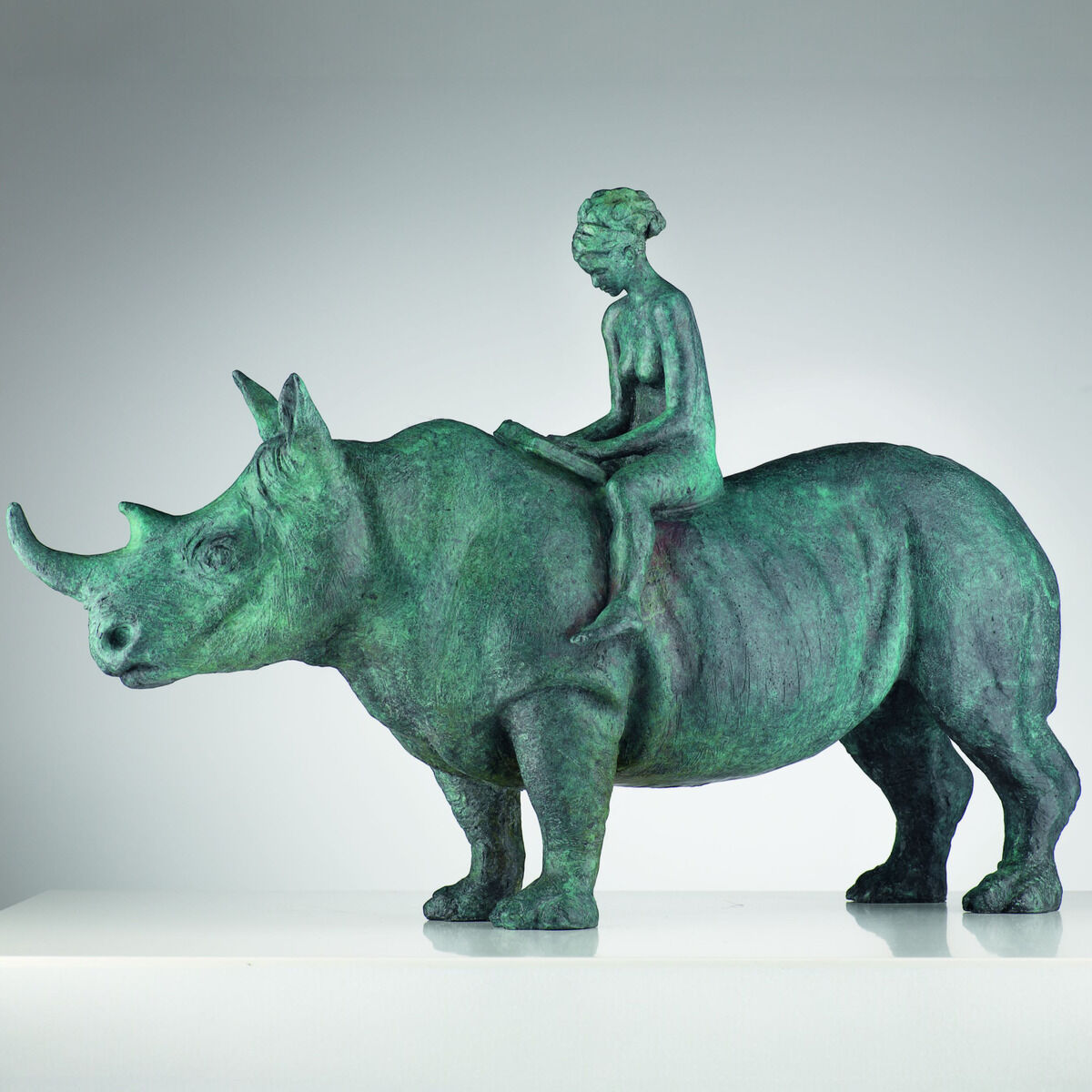 Auf weißem Sockel steht Alfredo Llorens Garcías grün patinierte Bronzeskulptur „The Maid and the Unicorn“: Ein massiges Nashorn im Profil nach links trägt auf seinem Rücken eine schlanke, aufrecht sitzende Frau mit aufgeschlagenem Buch. Die handziselierte Oberfläche zeigt plastische Hautfalten, Horn, Ohren und Beine; neutral grauer Hintergrund.