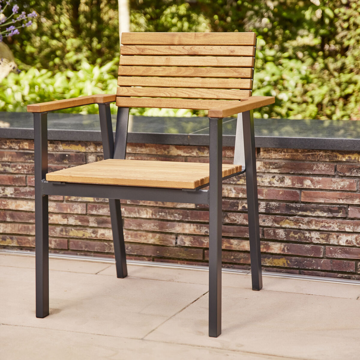 Ein Stapelsessel aus Holz und Metall steht auf einer Terrasse mit einem Hintergrund aus grünen Büschen und einer Backsteinmauer. Der Stuhl hat eine Holzsitzfläche und Armlehnen sowie ein schwarzes Metallgestell.