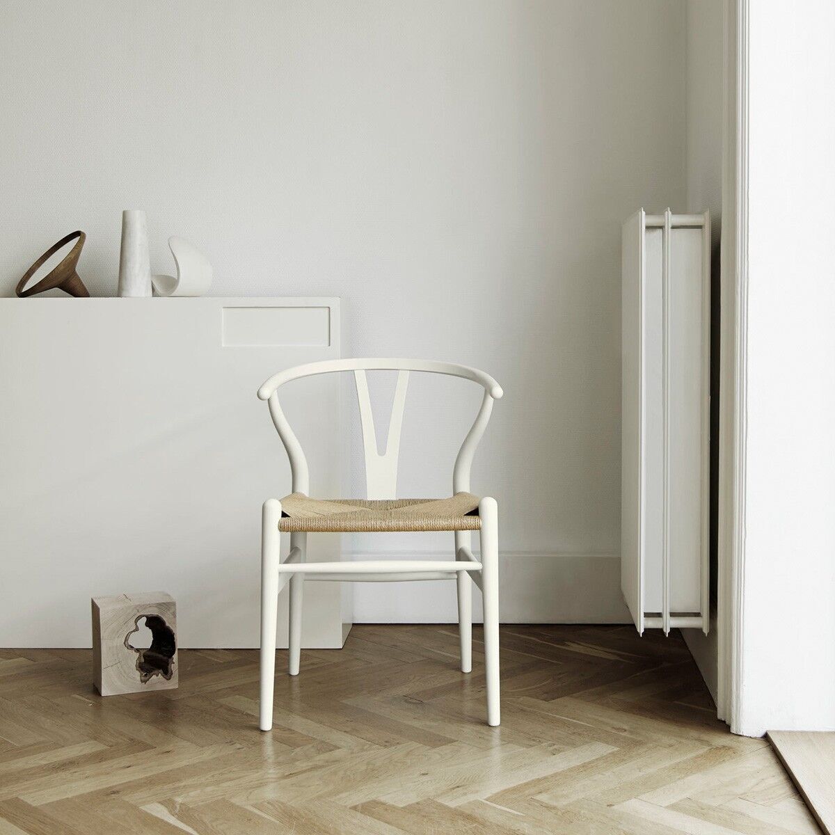 Sonderedition »Stuhl CH24 Soft« von Carl Hansen. Ein eleganter, weißer Holzstuhl in einem minimalistischen, skandinavischen Design, der vor einer weißen Wand und einem Heizkörper platziert ist. Daneben befindet sich ein minimalistisches Regal mit Dekoration.