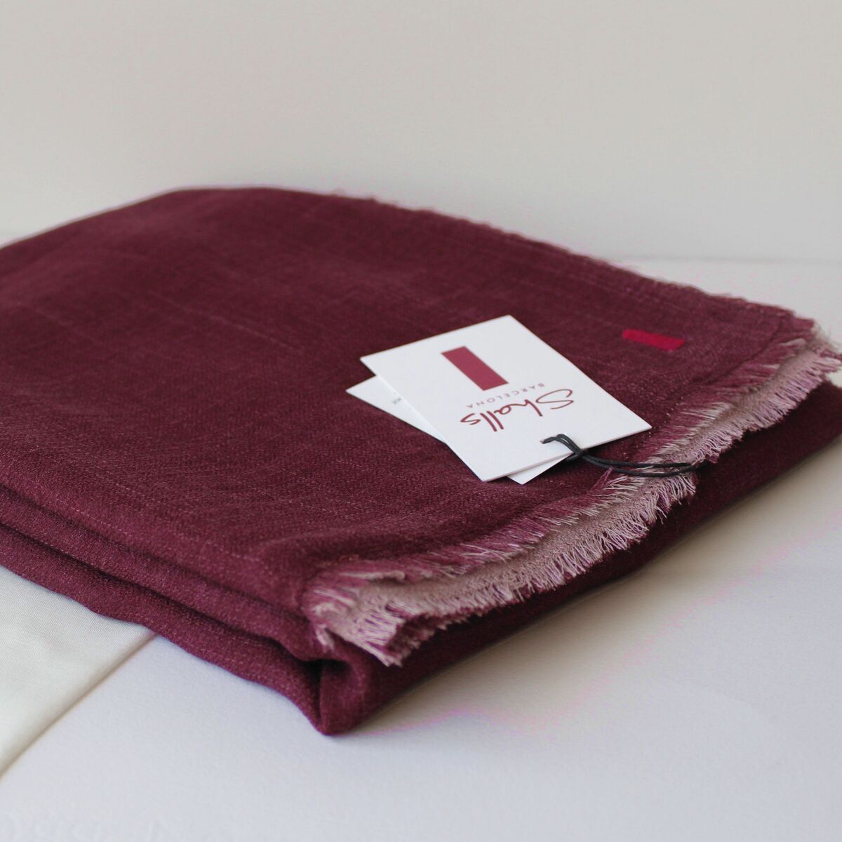 Gefalteter, weinroter Wollschal mit heller Rückseite und Fransenrand. Daneben liegt ein weißer Stoffbeutel und ein Anhänger mit der Aufschrift „Carla“. Das Produkt ist auf einem neutralen Hintergrund präsentiert.