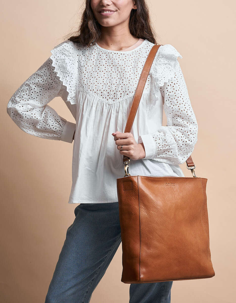 Eine Frau trägt eine braune Ledertasche »Sofia« über der Schulter. Sie trägt eine weiße, langärmelige, Spitzenbluse und eine blaue Jeans. Der Hintergrund ist beige.