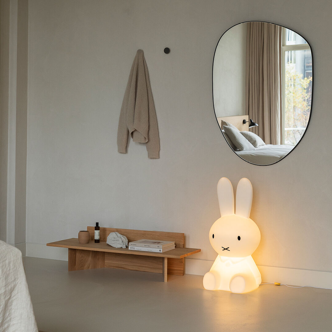 Im Bild ist eine freundlich aussehende, beleuchtete Lampe in Form einer bekannten Kinderfigur namens Miffy XL zu sehen. Die Lampe steht auf dem Boden eines modernen und minimalistisch eingerichteten Zimmers mit neutralen Farben. An der Wand hängt ein Spiegel und daneben ein Handtuch. Eine kleine Bank, auf der ebenfalls ein Buch und Dekorationsartikel stehen, verleiht dem Raum zusätzliche Behaglichkeit.