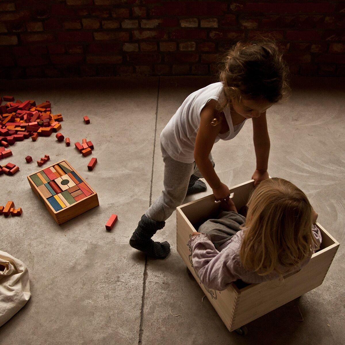 Zwei Kinder spielen mit bunten Holzbausteinen. Einige Bausteine sind noch auf dem Boden verteilt, während ein Kind in einer Holzkiste sitzt und das andere Kind sich davor bückt.