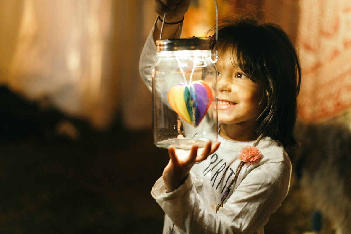 Ein Kind hält ein Sonnenglas Classic, eine nachhaltige Solarlampe, die in einem Einmachglas untergebracht ist. Im Glas hängt eine bunte Herzdekoration. Das Kind lächelt und trägt ein weißes Oberteil.