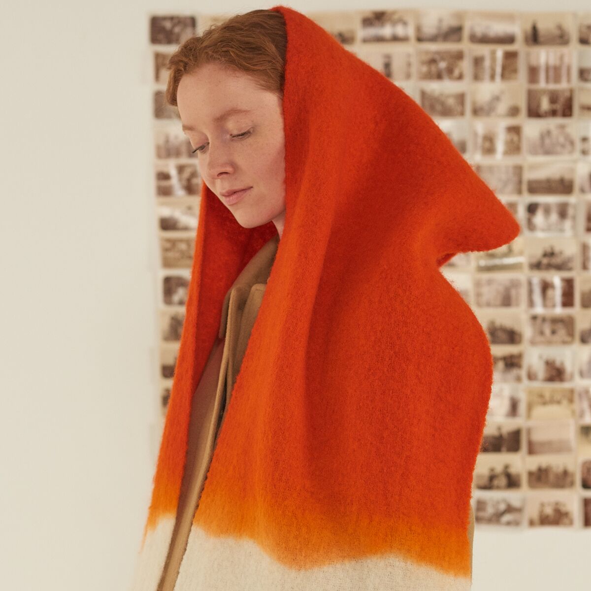 Eine Person trägt einen auffälligen Schal »No.698« von Moismont in der Farbvariante „tomato“, ein kräftiges Orange, das in Cremeweiß übergeht. Der Schal ist voluminös, aus Schurwolle und Nylon gefertigt und weich strukturiert. Unscharfer Hintergrund mit Sepia-Fotowand.