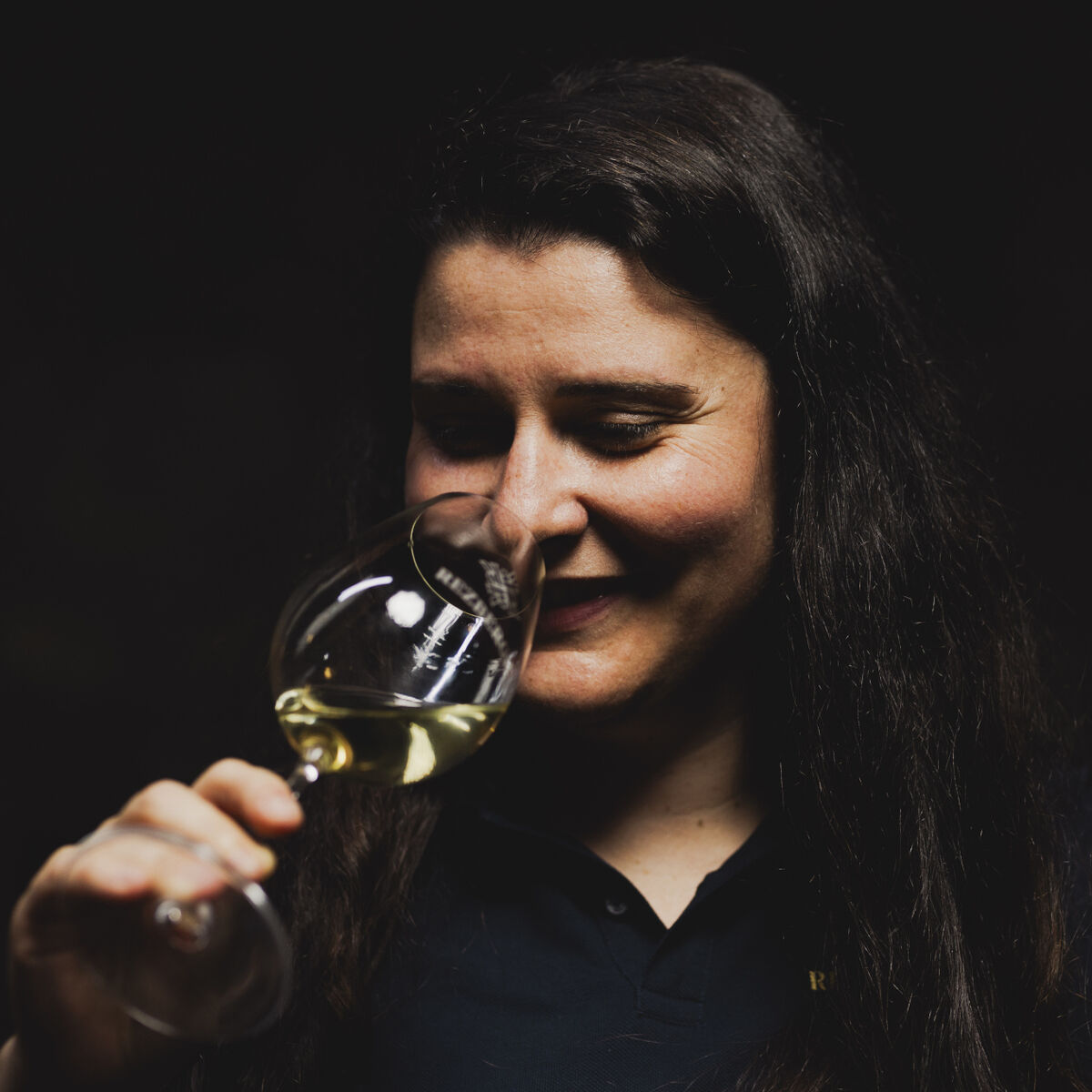 Eine Frau hält ein Glas mit Crémant de Luxembourg BRUT und lächelt, während sie den Schaumwein betrachtet. Die Szene ist stimmungsvoll und dunkel gehalten, der Fokus liegt auf dem Glas und ihrem Gesicht.