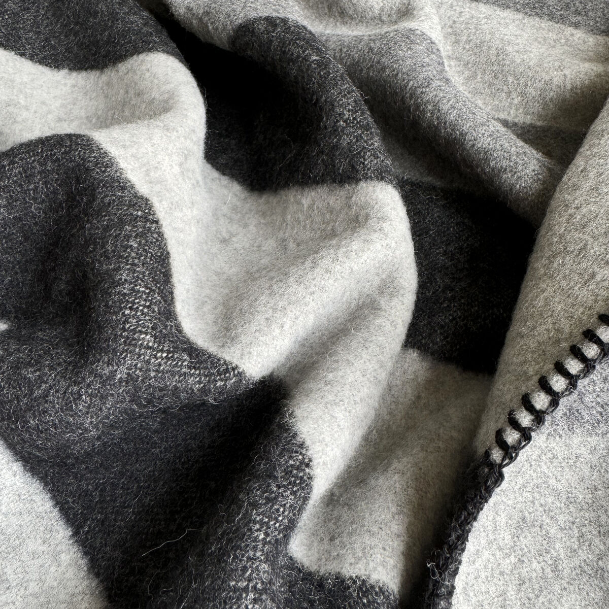 Nahaufnahme einer edlen Kaschmirdecke mit anthrazitfarbenen und hellgrauen Streifen, weich und flauschig mit dekorativer Kettelnaht am Rand, ideal als luxuriöses Wohnaccessoire für gemütliche Stunden.