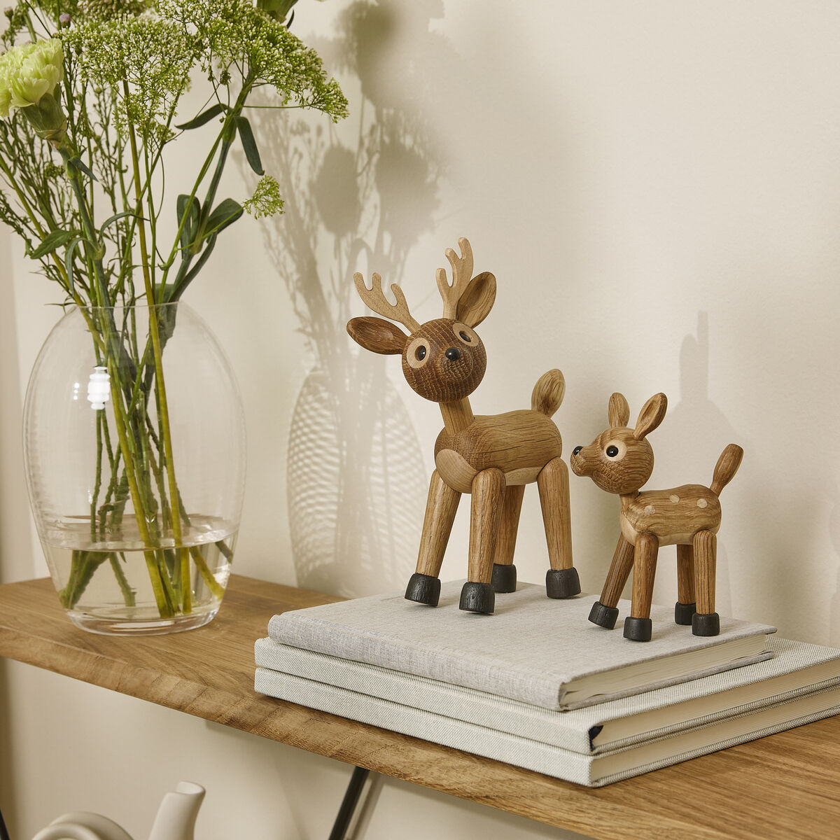 Zwei aus Holz gefertigte Rehfiguren stehen auf gestapelten Büchern auf einem Holzregal. Im Hintergrund befindet sich eine Glasvase mit frischen Blumen sowie weitere Dekoelemente. Die Szene wirkt modern und freundlich.