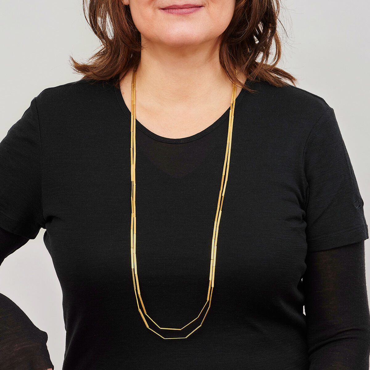 Eine Frau trägt eine lange, goldplattierte Kette namens »Streifen« über einem schwarzen Oberteil. Die Kette ist in minimalistischem Design gehalten und verleiht einen modernen, eleganten Look.