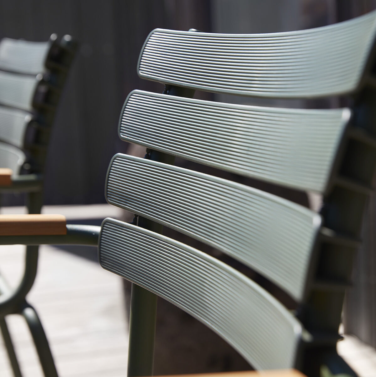 Nahaufnahme eines Stuhls »RECLIPS« mit grüner Sitzfläche und schwarzem Metallrahmen, der sich auf einer Terrasse befindet. Die Armlehne des Stuhls besteht aus Holz.