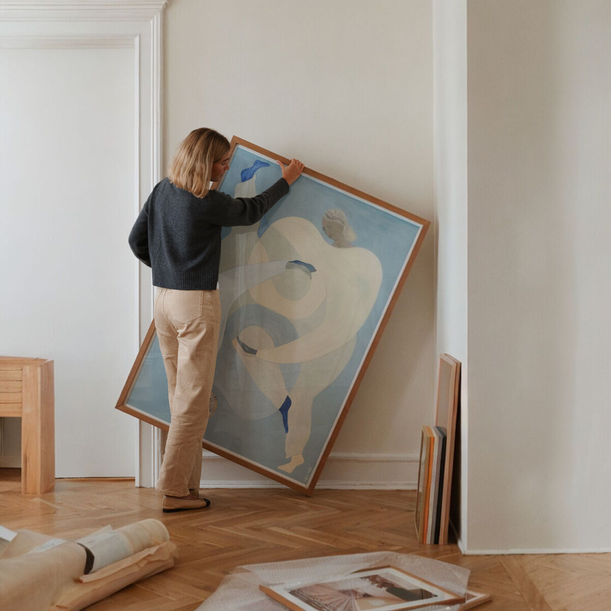 Eine Frau in einer Wohnung arrangiert ein großes gerahmtes Gemälde mit abstrakten, blauen und beigen Formen an einer hellen Wand.