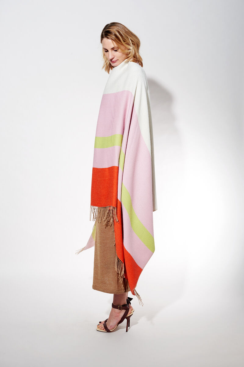 Eine Frau trägt ein Cape aus Alpaka-Wolle mit den Farben Weiß, Rosa, Grün und Orange. Das Cape hat Fransen und bietet eine stilvolle und warme Ergänzung zu ihrem Outfit.