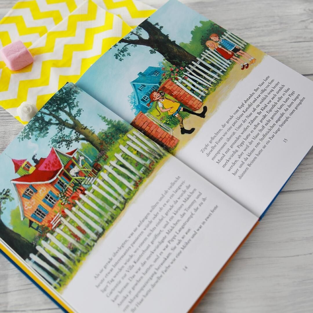 Farbiges Kinderbuch 'Pippi Langstrumpf' mit Illustrationen, die Pippi neben einem weißen Zaun und vor einem roten Haus zeigen. Das Buch liegt auf einer weißen Holzoberfläche neben einem gelb-weiß gemusterten Stück Stoff.