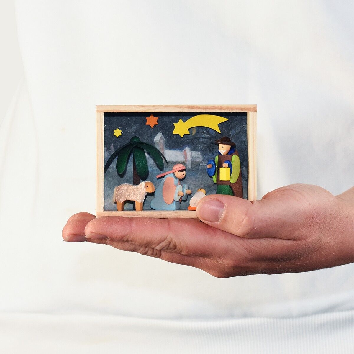 Ein kleines Holzfiguren-Set zeigt die Weihnachtskrippenszene mit Maria, Josef, dem Jesuskind, einem Schaf, einem Stern und einem Engel. Das Set ist in einer Holzkiste auf einer Hand präsentiert.