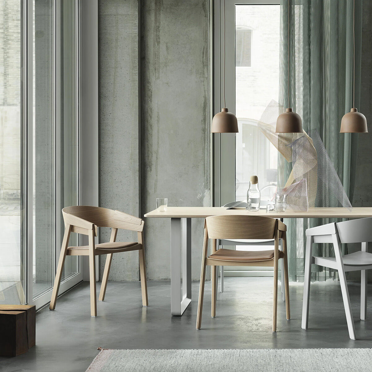 Helles, modern eingerichtetes Zimmer mit großem Fenster, grauem Sichtbeton, hellen Eichenholzstühlen vom Typ Muuto Cover Stuhl »Eiche« an einem schlichten weißen Tisch, dekoriert mit Glasflaschen und zwei kupferfarbenen Hängelampen im skandinavischen Stil.