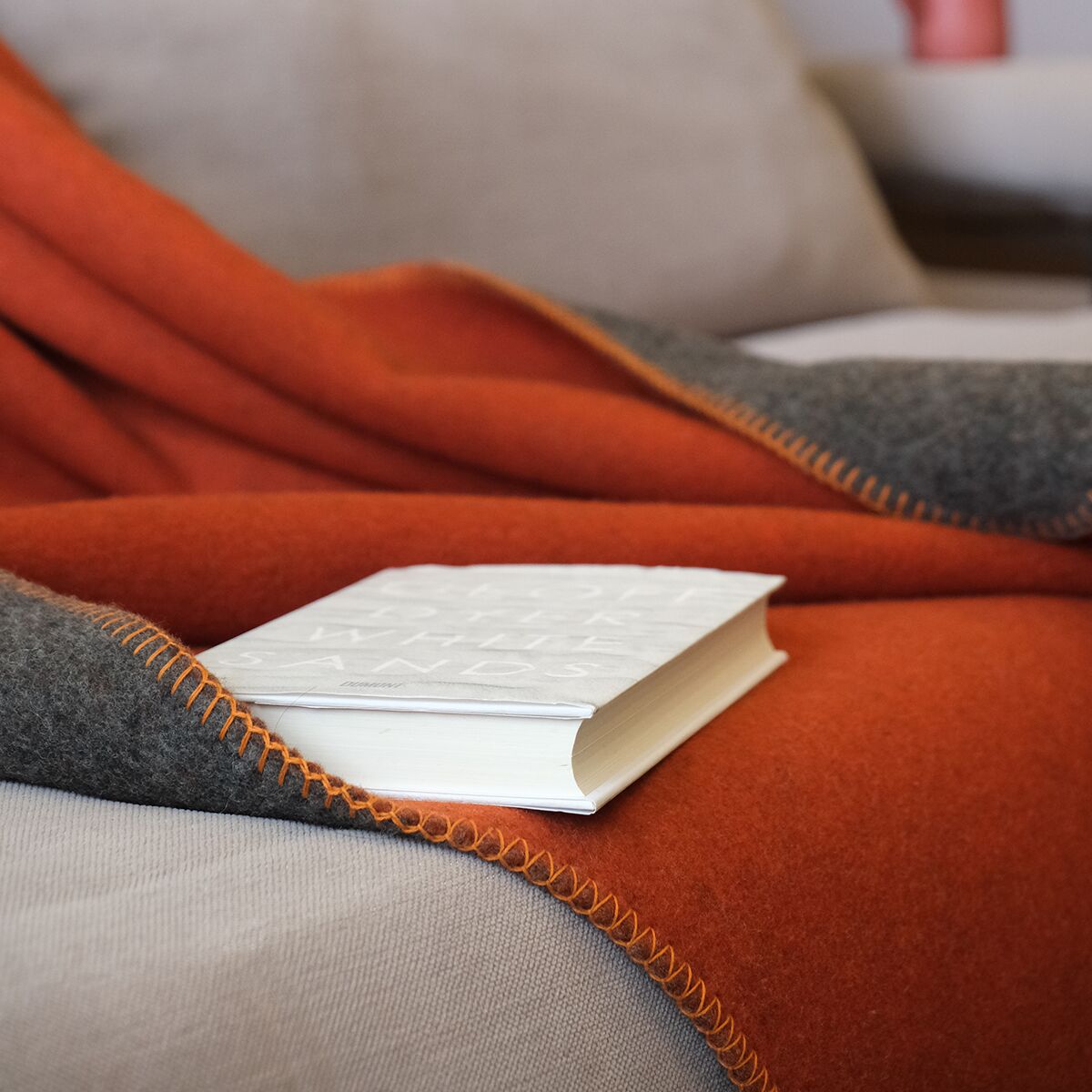 Eine orange und graue Merinodecke namens »FireSideMoon«, die auf einem beigen Sofa liegt. Auf der Decke befindet sich ein weißes Buch.