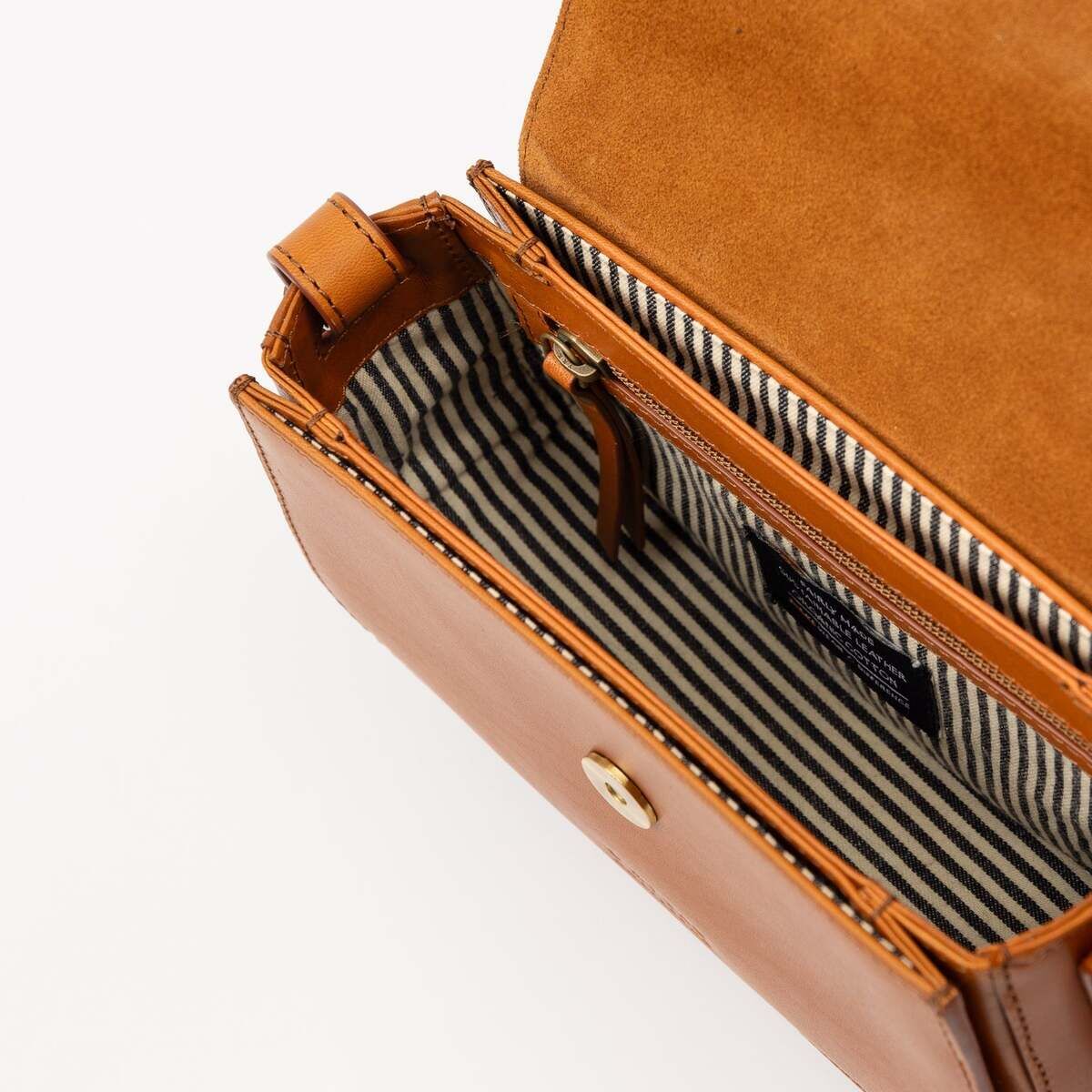 Offene, braune Ledertasche ‚Kendalini‘ mit gestreiftem Innenfutter, sichtbarem Reißverschlussfach und Magnetverschluss, fotografiert auf hellem Hintergrund.
