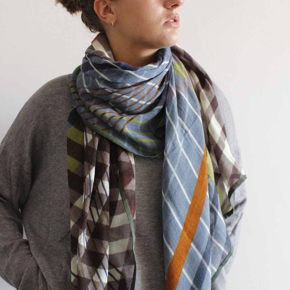 Frau mit einem groß gemusterten Wollschal in Mauvetönen, Blau, Braun, Grün und Orange, locker um den Hals gelegt. Sie trägt einen grauen Pullover, das Bild wirkt herbstlich und gemütlich. Das Foto zeigt das stilvolle Accessoire als modisches Highlight.