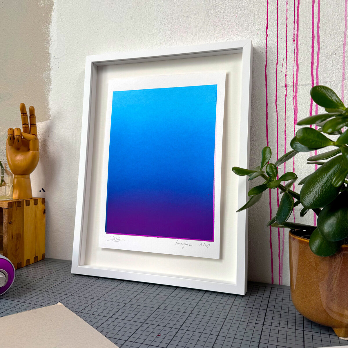 Gerahmtes Kunstwerk von Daniel Lisson, Titel »Imagine« (Blue/Violet), im Format 40x30 cm. Das Motiv zeigt einen sanften Farbverlauf von kräftigem Blau zu Violett, gerahmt in weißem Eichenholz, angelehnt an eine Wand. Im Vordergrund stehen eine hölzerne Hand, eine Pflanze und weitere Deko-Elemente.