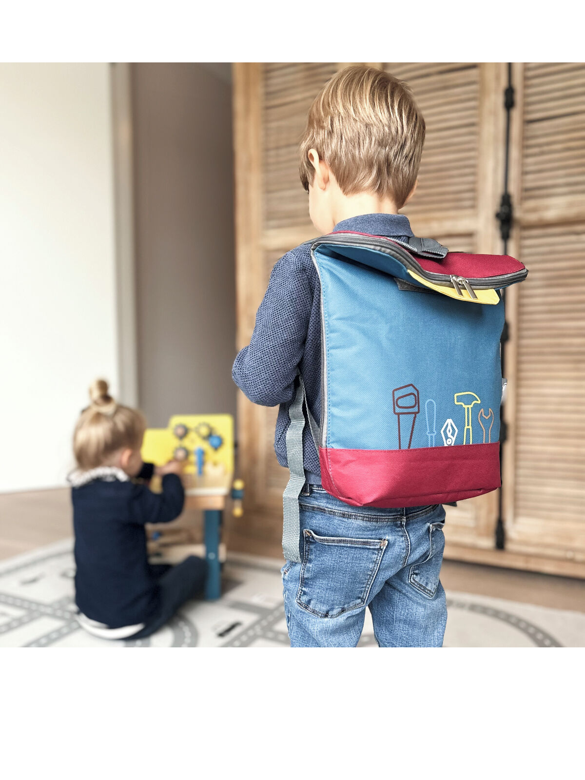 Ein Kind mit einem blauen und bordeauxroten Werkzeugrucksack steht auf einem Spielteppich, während ein weiteres Kind am Werkbank-Spielzeug arbeitet. Im Hintergrund ist eine Holztür zu sehen.