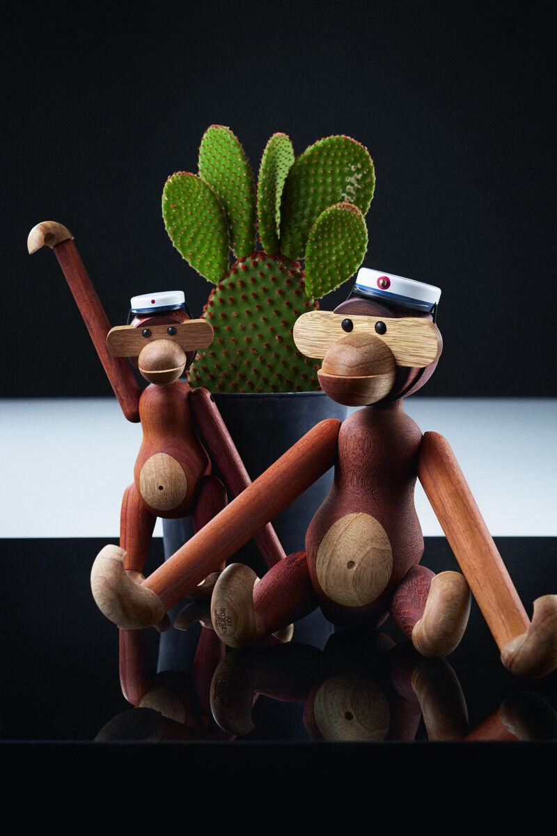Zwei Holzfiguren in Form von Affen von Kay Bojesen, eine stehend und eine sitzend, vor einem Kaktus auf einem schwarzen Hintergrund. Die Figuren tragen Mützen mit weißem und schwarzem Band und rotem Knopf.